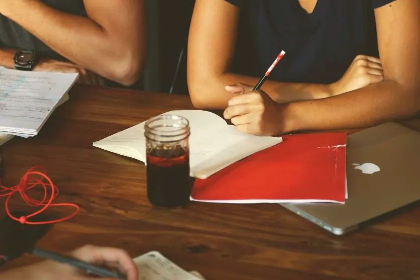 Dos personas toman notas en una mesa de madera con un cuaderno, una carpeta roja y un portátil